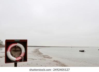 Lifebuoy in a wooden box on a deserted beach with a calm bay and boats in the distance, under an overcast sky - Powered by Shutterstock