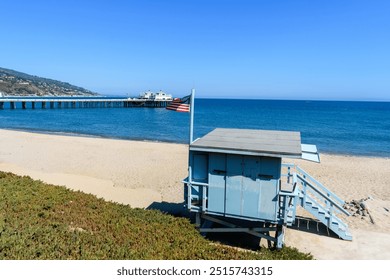 A lifeguard tower on a sandy beach in Malibu with Malibu Pier and calm ocean in the background, under a clear blue sky - Powered by Shutterstock