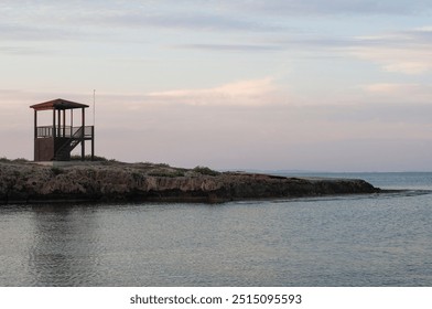 A lifeguard tower stands alone on a rocky outcrop, offering a view of the tranquil sea under a pastel sky at twilight, creating a peaceful coastal atmosphere. - Powered by Shutterstock