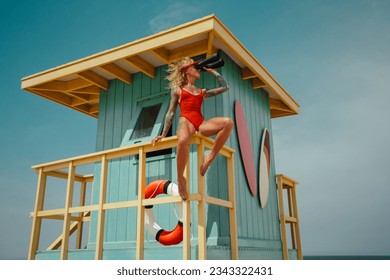 Lifeguard woman in red bikini and visor with binocular and lifebuoy on watch tower against turquoise sky