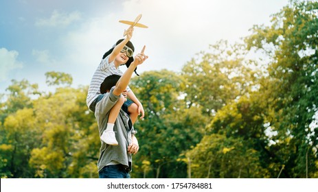 Lifestyle Asian Family Father And Son Sat On His Shoulders And Run In  Park. Paper Plane As A Toy In Hand Of Kid And There Are Many Big Trees In Garden Is Natural In The Morning Of Summer