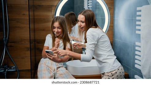 Lifestyle. Beautiful caucasian girls brush their teeth and read messages on phone from bathroom at home. Two girls brushing their teeth and use smartphone, checking social networks - Powered by Shutterstock