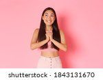 Lifestyle, beauty and women concept. Portrait of dreamy beautiful asian girl, smiling pleased as have wish, clasp hands together in pray, looking upper left corner while pleading, pink background