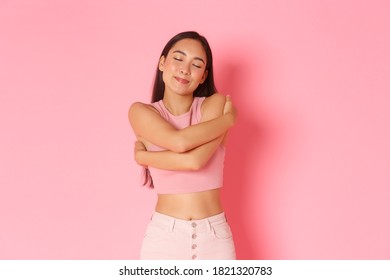 Lifestyle, beauty and women concept. Portrait of carefree, romantic asian girl smiling while daydreaming, close eyes and remember moment, hugging herself, standing over pink background