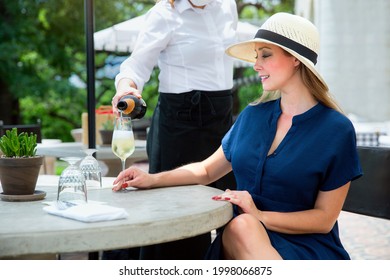 Lifestyle candid portrait of a server pouring sparkling wine to an elegant and classy woman, seated outdoors on countryside restaurant patio on a beautiful summer day