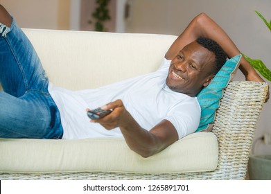 Lifestyle Candid Portrait Of Young Attractive And Happy Black African American Man Holding TV Remote Controller Watching Television Movie Enjoying Relaxed Chilling At Living Room Lying At Home Couch