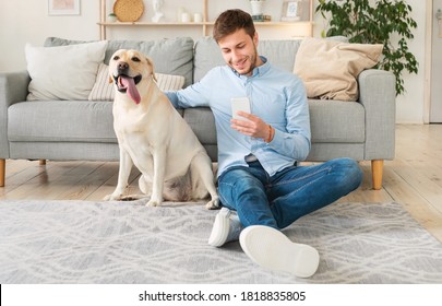 Lifestyle Concept. Portrait of smiling man using smartphone sitting on the carpet with his labrador, stroking dog. Guy spending time at home, chatting at social media, texting sms or reading message