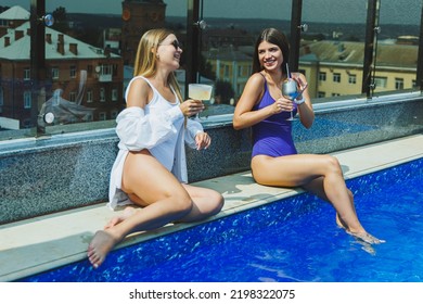 Lifestyle Concept. Two Attractive Women In Sunglasses Drinking Cocktails By The Pool. Side View Of Glamorous Hot Ladies In Swimsuits Having Fun In Turquoise Pool.