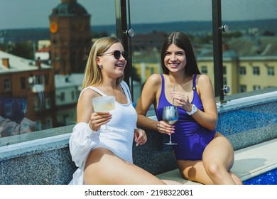 Lifestyle Concept. Two Attractive Women In Sunglasses Drinking Cocktails By The Pool. Side View Of Glamorous Hot Ladies In Swimsuits Having Fun In Turquoise Pool.