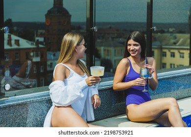 Lifestyle Concept. Two Attractive Women In Sunglasses Drinking Cocktails By The Pool. Side View Of Glamorous Hot Ladies In Swimsuits Having Fun In Turquoise Pool.