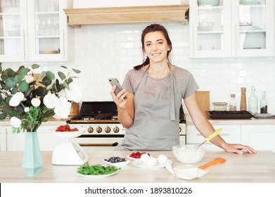 Lifestyle, Cooking And Freelance Concept: Young Brunette Woman Looking At A Recipe On Her Smartphone And Getting Ready To Make A Cake