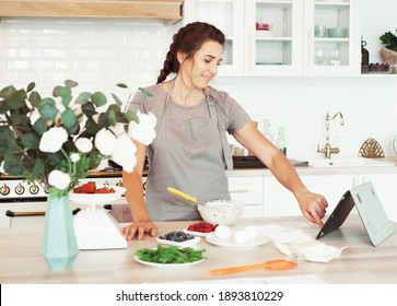 Lifestyle, Cooking And Freelance Concept: Young Brunette Woman Looking At A Recipe On Her Tablet And Getting Ready To Make A Cake