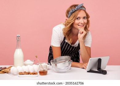 Lifestyle, Cooking. Young Adult Blonde Woman Looking At Camera And Holding Chin, Reading Recipe On Her Tablet And Getting Ready To Make Cake. Indoor Studio Shot Isolated On Pink Background.