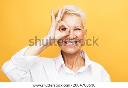 lifestyle, emotion and old people concept: Senior grey-haired woman smiling happy doing ok sign with hand on eye looking through fingers over yellow background