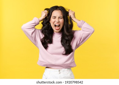 Lifestyle, emotions and advertisement concept. Angry distressed asian woman screaming hateful and outraged, feeling pissed-off and fed up, pulling her hair with anger and rage, yellow background