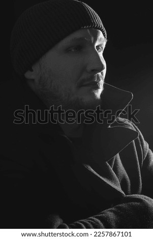 Lifestyle, fashion, occupation concept. Man with winter hat and black coat studio portrait. Model with beard looking aside the camera with serious look. Dark studio background. Black and white image