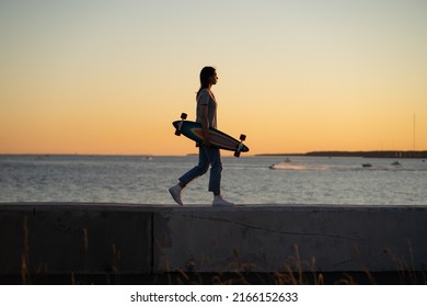 Lifestyle and freedom concept: young girl with longboard walking over sunset view. Silhouette of trendy teen or student female skater with skateboard at riverside pavement. Woman walk along river