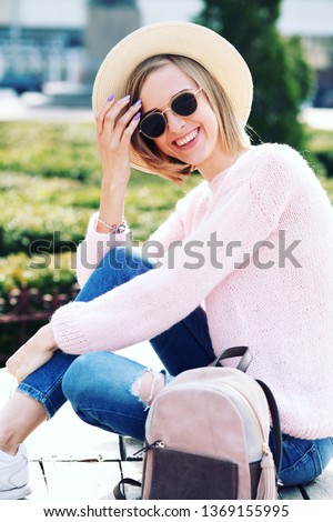 Lifestyle Image of carefree traveling smiling woman walking in spring city. Hipster outfit. Oversize pink sweater, straw hat, round sunglasses and stylish accessories.