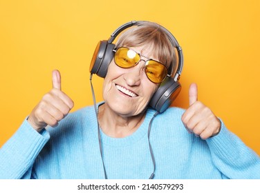 Lifestyle and people concept: Funny old lady listening music and showing thumbs up. Isolated on yellow background.