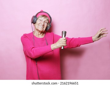 lifestyle and people concept: Funny old lady listening music with headphones and singing with mic over pink background