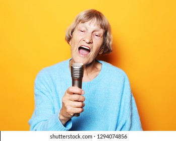 Lifestyle  And People Concept: Happy Old Senior Woman Singing With Microphone, Having Fun, Expressing Musical Talent Over Yellow Background