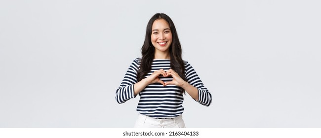 Lifestyle, People Emotions And Casual Concept. Lovely Smiling Adorable Asian Woman Showing Heart Sign And Smiling, Express Sympathy Or Care, Standing White Background