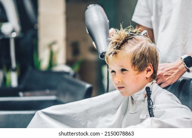 Lifestyle Photo Barber Shop Blond Boy Customer. Hairdresser Man Work Move Hand Comb Tool Scissors Dryer Clip White Cape. Modern Style Kid Short Hair Cut. Fashion Business Service For Children