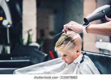 Lifestyle Photo Barber Shop Blond Boy Customer. Hairdresser Man Work Move Hand Comb Tool Scissors Dryer Clip White Cape. Modern Style Kid Short Hair Cut. Fashion Business Service For Children