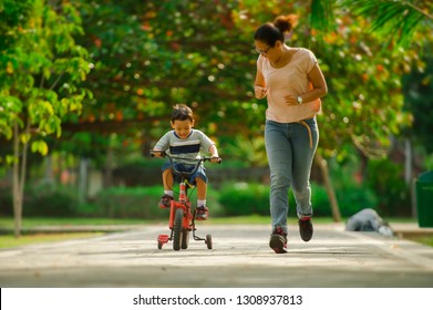 Lifestyle Portrait Of Asian Indonesian Mother And Young Happy Son At City Park Having Fun Together The Kid Learning Bike Riding And The Woman Running After The Child In Motherhood Concept