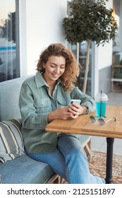 Lifestyle Portrait Of Beautiful Millennial Woman 25-30 Years Old With Curly Hair Sitting In Cafe And Laughing, Using Mobile Phone, Texting Message 