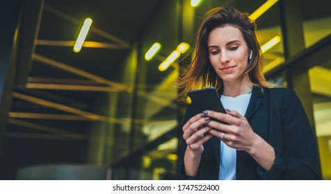 Lifestyle Portrait Business Woman Walking In Evening City And Using Mobile Gadget, Girl Banker Dressed In Business Style Listen To Music Wireless Earphones And Typing Text Message On Smartphone Device