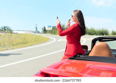 Lifestyle portrait of a carefree beautiful woman in sunglasses and red suit standing near red cabriolet car applying lipstick. Road trip enjoying freedom, business lady lifestyle concept  - Powered by Shutterstock