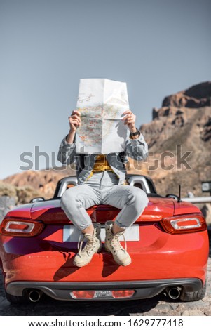 Lifestyle portrait of a carefree woman dressed casually in jeans and red hat sitting with map on the car trunk, enjoying road trip on the desert valley
