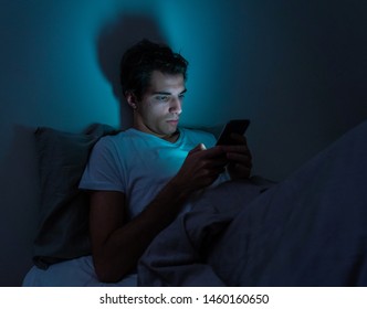 Lifestyle Portrait Of Cell Phone Addict Man Awake Late At Night In Bed Using Smart Phone Checking Likes And Followers On Social Media. Internet Addiction And Mobile Abuse.