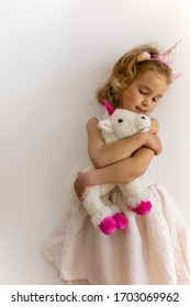Lifestyle portrait of cute positive happy little toddler girl wearing pink unicorn tiara in white ball dress gown hugging stuffed soft unicorn toy isolated on white background cutout with copy space.