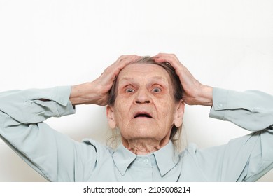 Lifestyle Portrait Of An Elderly Woman Holding Her Hands In Her Hair On Her Head On A White Background. Surprised Old Pensioner With A Frightened Expression Face. Concept Of Emotional Shock.