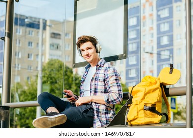 Lifestyle portrait of hipster student man with smart phone sitting on bench with yellow bagpack and phone on the tram stop in the modern city.
