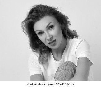 Lifestyle Portrait Of Middle Aged Caucasian Blonde Woman In White T-shirt And Blue Jeans Sitting On Floor In Bright Interior Near White Wall. Barefoot Female