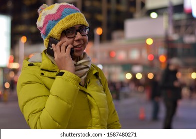 Lifestyle Portrait Shot Of Smiling Young Indian Woman, Wearing Jacket And Cap, Speaking On Her Mobile Phone In The American Winters. Bokeh Of Christmas Lighting Can Be Seen As Boken In The Background