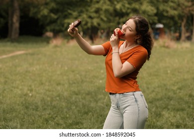 Lifestyle Portrait Of Smiling Curly Girl College Student Eating Red Juicy Apple And Holding Ice Lolly Cream In Hand. Pretty Woman Lead Healthy Lifestyle In The City Park With Blur Background.