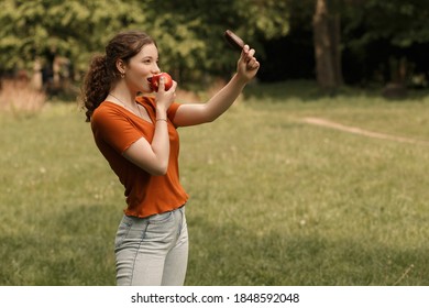 Lifestyle Portrait Of Smiling Curly Girl College Student Eating Red Juicy Apple And Holding Ice Lolly Cream In Hand. Pretty Woman Lead Healthy Lifestyle In The City Park With Blur Background.