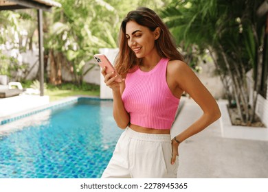 Lifestyle portrait of smiling woman using mobyle phone .  Posing in modern villa with pool.  Wearing stylish pink crop top and white pants. - Powered by Shutterstock