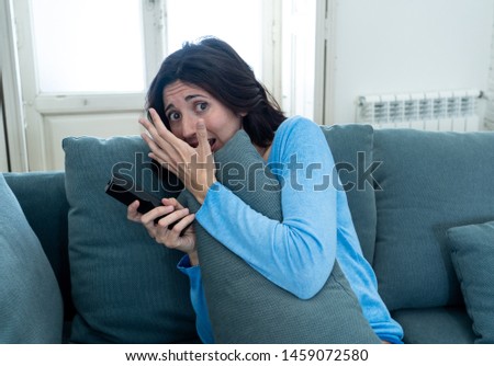 Lifestyle portrait of woman feeling scared and shocked making fear, anxiety gestures while watching horror movie on TV holding remote control. In horror and violence on TV and internet and mass media.