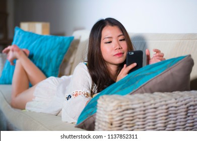 lifestyle portrait of young beautiful and happy Asian Korean woman on her 20s or 30s lying at living room sofa couch using internet mobile phone looking gorgeous and sweet in communication