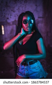 Lifestyle, Portrait Of A Young Black Woman With Long Braids, Sunglasses And A Black T-shirt. Pink And Blue Neon Lights, Urban Photography Of A Trap Dancer