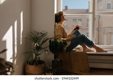 Lifestyle portrait of young brunette sitting on windowsill in light room. Cute short-haired lady wearing orange shirt and blue jeans resting, holding cup of coffee and phone near green