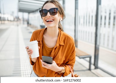 Lifestyle portrait of a young and cheerful woman standing with phone and coffee cup on the public transport stop outdoors. Urban business travel and transportation concept