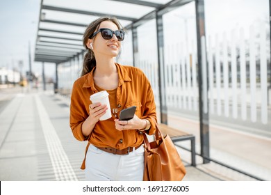 Lifestyle Portrait Of A Young And Cheerful Woman Standing With Phone And Coffee Cup On The Public Transport Stop Outdoors. Urban Business Travel And Transportation Concept