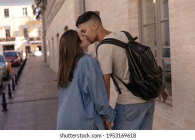 Lifestyle Portrait Of Young Couple Walking On Street And Kissing. Cute Dark-haired Boy Wearing Beige T-shirt And Black Backpack Posing With Pretty Girl In Denim Shirt