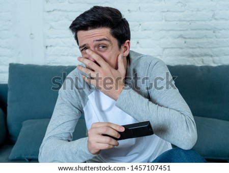 Lifestyle portrait of young man feeling scared and shocked making fear, anxiety gestures while watching television holding remote control. In horror and violence on TV and internet and mass media.